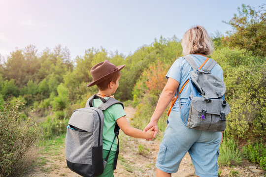 mother and son with backpacks holding hands hiking on forest trail back view