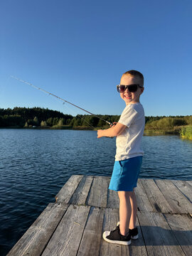 Little boy in sunglasses holding a fishing rod and fishing on the lake. Child standing on a wooden pier outdoors in nature.