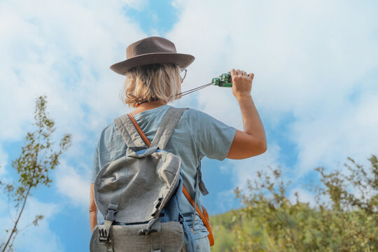 woman tourist with hat and backpack looking through binoculars in nature on sunny summer day