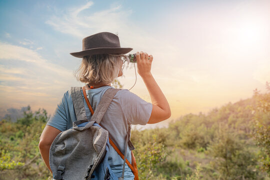woman tourist with hat and backpack looking through binoculars in nature on sunny summer day