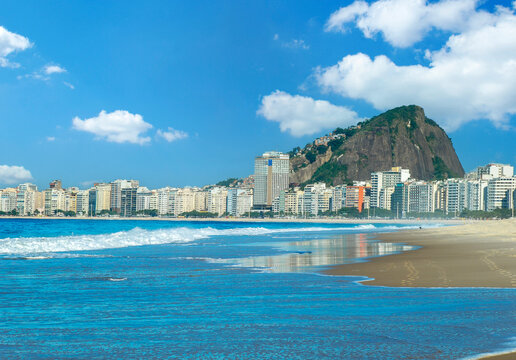 Scenic daytime view of Copacabana beach in Rio de Janeiro, Brazil, featuring vibrant blue ocean waves, sandy shoreline, city skyline, and mountains under a sunny blue sky.