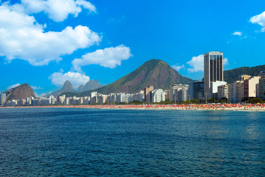 Panoramic view of Copacabana beach in Rio de Janeiro, Brazil. Features crashing ocean waves, the city skyline, and iconic mountains under a bright, sunny blue sky with clouds.