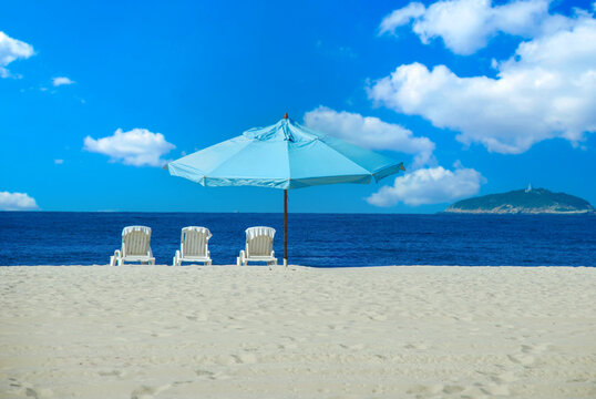 Beautiful sunny day at Copacabana beach in Rio de Janeiro with three white lounge chairs and a blue umbrella on the white sand, facing the blue ocean.