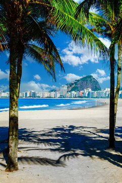 Scenic view of Copacabana beach, Rio de Janeiro, framed by palm trees on white sand, featuring the blue ocean, city skyline, and mountains under a sunny sky.