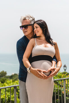 Expecting couple embraces outdoors while forming a heart with their hands over a pregnant belly. Sunny maternity portrait conveys love, family, anticipation, and new beginnings.