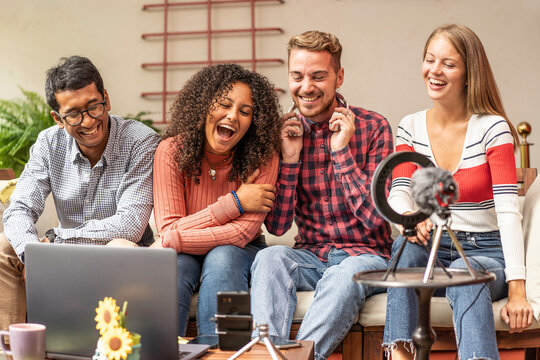 Multiethnic gen z creators laugh while recording a group podcast and livestream in a home studio. A laptop, camera, ring light, microphone, and headphones frame collaborative digital content.