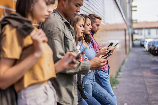 Multiethnic gen z friends stand together while absorbed in smartphones beside an urban wall during an outdoor meetup. Candid street scene reflects phubbing, screen dependence, and social disconnection