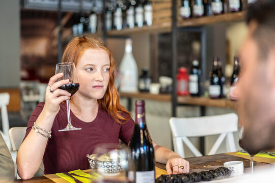 Serious gen z woman holds a glass of red wine while looking toward a companion during an indoor tasting. Candid scene suggests moderation, hesitation, social pressure, and responsible drinking.