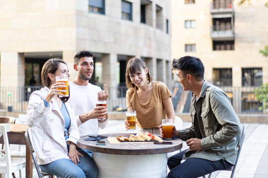 Young friends laughing as a man raises his index finger during outdoor drinks. Multiethnic millennials and gen z share beer, jokes and lively conversation at a city cafe.