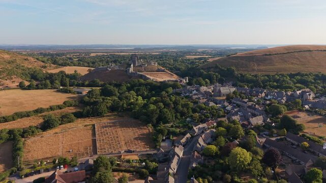 Aerial drone footage of the iconic Corfe Castle in Dorset, England, showcasing the medieval castle ruins, historic village, and rolling Purbeck countryside on a hot summer morning
