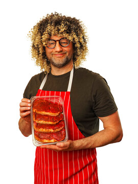 Man in his 40s or 50s with curly hair holds tray of marinated strip loin steaks. He wears red and white striped apron. Background is plain white. Funny looking butcher promoting his craft product.