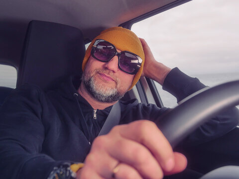 Male driver in his 40s or 50s in dark clothes and yellow hat and dark sun glasses. Man with grey beard behind driving wheel, happy friendly face Transport and tourism.