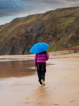 Raindrops patter on a vivid blue umbrella. Figure in purple puffer jacket stands alone. Wet sand reflects the gray, stormy sky. Cliffs loom in the background, wild and imposing. Travel and tourism.