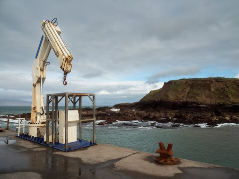Crane lifts metal structure near rocky coastline. Weathered pier with rusted bollard. Ocean waves crash against dark cliffs. Grey clouds cover sky. Concrete platform wet from sea spray