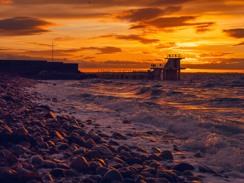 Ocean rough rock shoreline and silhouette of Blackrock diving board in Salthill, Galway city, Ireland. Warm orange sunrise sky in the background. Town landmark and tourist attraction.
