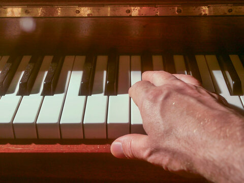 Hand on piano keys under warm, soft lighting. Shallow depth of field highlights keyboard texture. Wooden casing partially visible in background. Focus on black and white keys with gentle blur
