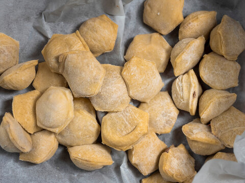 Freshly made, uncooked dumplings on parchment paper. Golden brown, irregularly shaped dumplings scattered across surface. Ready for cooking or baking, homemade style. Textured dough visible