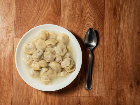 White bowl filled with dumplings in broth, placed on wooden table. Metal spoon rests beside the bowl. Simple, rustic meal presentation with warm lighting. Popular Russian dish. Fast food. Simple meal.