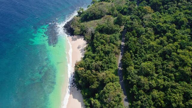 Aerial view of Waebela beach with turquoise water, sandy shore, and lush forest bordering the coastal road in Inerie, East Nusa Tenggara, Indonesia.