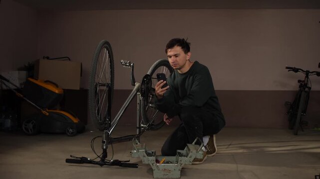 Man squatting in a garage looking at a smartphone while working on an upturned bicycle with a toolbox nearby, learning bike repair