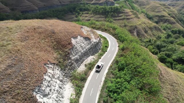 White car driving along a winding road through the rolling limestone hills of Sumba, Indonesia, surrounded by dry grasslands, lush valleys, and breathtaking tropical landscapes under a clear sky.