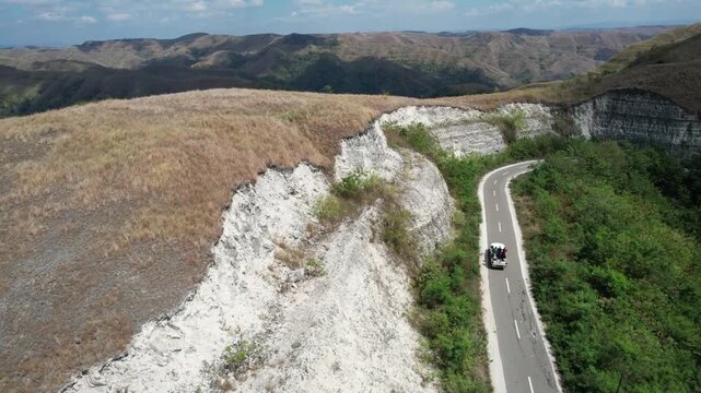 White car follows a winding mountain road beneath dramatic limestone cliffs and rolling hills in Sumba, Indonesia, showcasing rugged landscapes, tropical greenery, and scenic travel adventure.