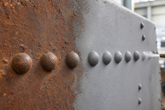 Sandblaster cleaning rust from steel girder surface with rivets. Industrial metal surface preparation using abrasive blasting process. Restoration, maintenance and construction metal work.