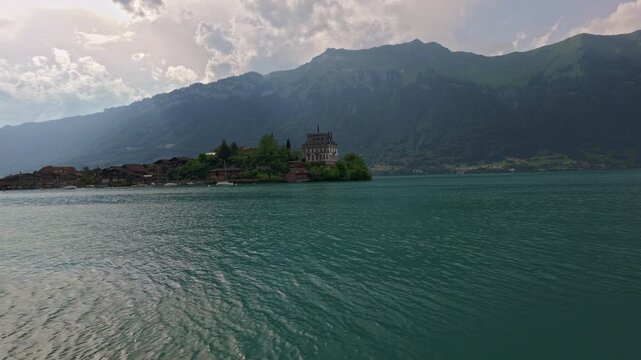 Slow pan over turquoise Lake Brienz toward Iseltwald peninsula with Seeburg Castle, alpine village, and dramatic Swiss mountains under cloudy sky. 4K UHD video.