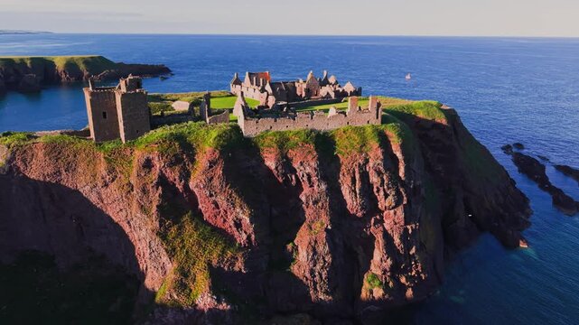 Aerial flight around Dunnottar Castle ruins on a dramatic sea cliff at sunset, overlooking the blue North Sea and rugged Aberdeenshire coast, Scotland. 4K UHD video.