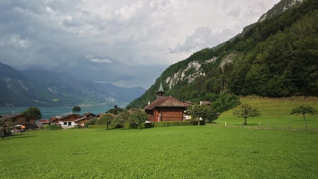 Pan around Iseltwald church amid green alpine meadows above Lake Brienz, with Swiss chalets, forested cliffs, and cloudy Bernese Oberland mountains, Switzerland. 4K UHD video.