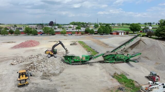 A large area of land is being transformed as workers operate machinery to crush and sort materials. Heavy trucks move in and out, carrying loads.
