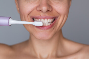 Fototapeta premium Close-up of young woman brushing teeth with electric toothbrush, smiling during morning oral hygiene routine on gray background