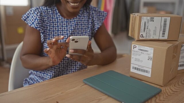 Woman smiling and tapping smartphone with hands beside stacked priority mail boxes on wooden desk in studio; entrepreneurial joy.