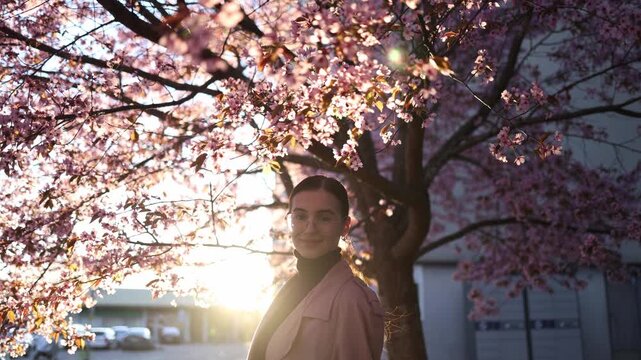 dark haired young woman standing near blooming cherry blossoms at sunset with warm evening light creating a peaceful spring lifestyle scene surrounded by delicate flowers and nature