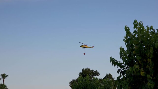 Firefighting helicopter carrying Bambi bucket with water to extinguish wildfire, slow motion video against clear blue sky with trees