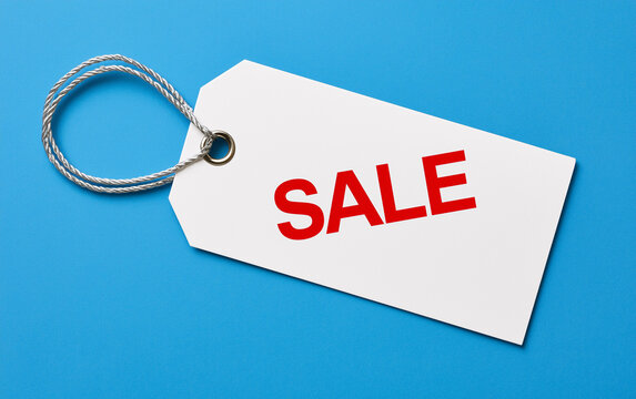 White paper price tag with red text sale word on blue background. Hanging label with string for retail commerce discount event.