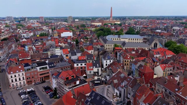 Aerial view video clip of the dense red roofed cityscape of Ghent in Belgium featuring parking lots, residential houses, modern developments, and industrial chimney on a bright daylight horizon