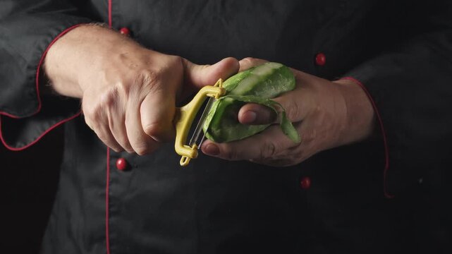 A chef in a black uniform skillfully peels a green cucumber with a yellow vegetable peeler, demonstrating the smooth removal of the skin while exposing the fresh green center in a well-lit kitchen