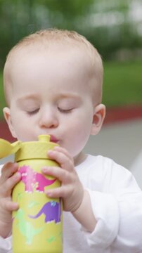 Caucasian toddler in white shirt holds yellow sippy cup, drinking from it while sitting outdoors in a green park, showing curiosity and engagement with surroundings