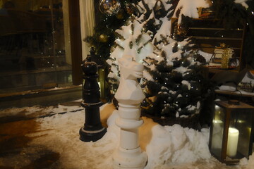 Large black and white chess knights stand on snowy ground near decorated pine trees and lit lanterns during a festive winter evening outside a shop window display © vannet