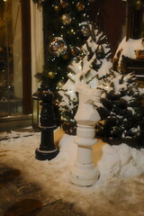Large black and white chess knights sit outside on a snowy sidewalk at night near glowing evergreen trees decorated with gold and silver ornaments for the holiday season © vannet