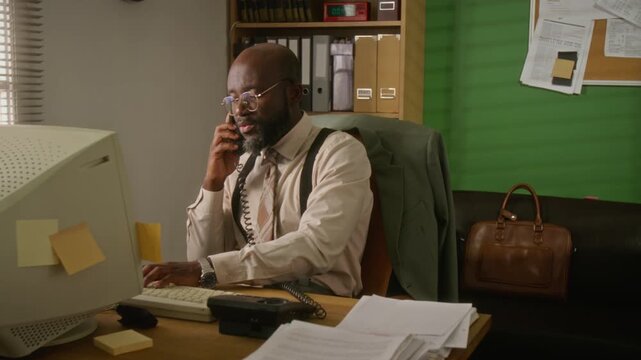 African American man talking on corded telephone and typing on vintage computer keyboard in retro office. Footage is perfect for nostalgic corporate training or retro tech marketing