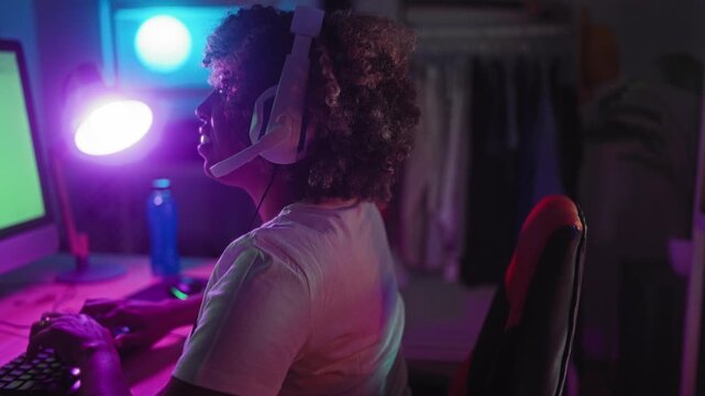 Woman with headset holding purple lamp and smiling while typing at gaming desk with keyboard, monitor, water bottle and red gaming chair in studio; joyful play.