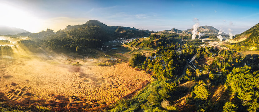 Aerial view of Dieng Plateau with volcanic steam rising from fumaroles over agricultural fields and rolling hills during golden hour in Indonesia.