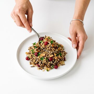 Close up of a serving spoon scooping wild rice pilaf with cranberries and almonds onto a white plate