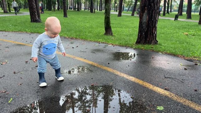 Baby boy explores puddles in a park after rain in rubber boots. 4k, slow motion.