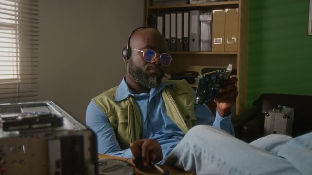 African American man wearing headphones examining and repairing computer graphics card with screwdriver. Perfect for IT support services and hardware repair tutorials