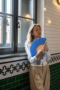 Woman feeling unwell suffering from heatstroke indoors, stands near window waving paper fan in hand to cool down, tired female with fatigue and dizziness caused by lack of fresh air during hot season.