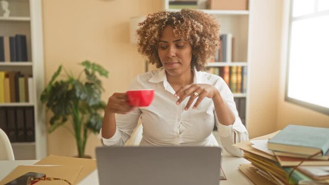 Woman typing on laptop at a cluttered desk with coffee cup and stack of books in a home study building, reaching for mug while working; focused productivity.