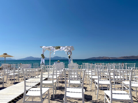 Elegant outdoor beach wedding ceremony setup with white chairs and floral arch overlooking turquoise Mediterranean sea under bright blue sky